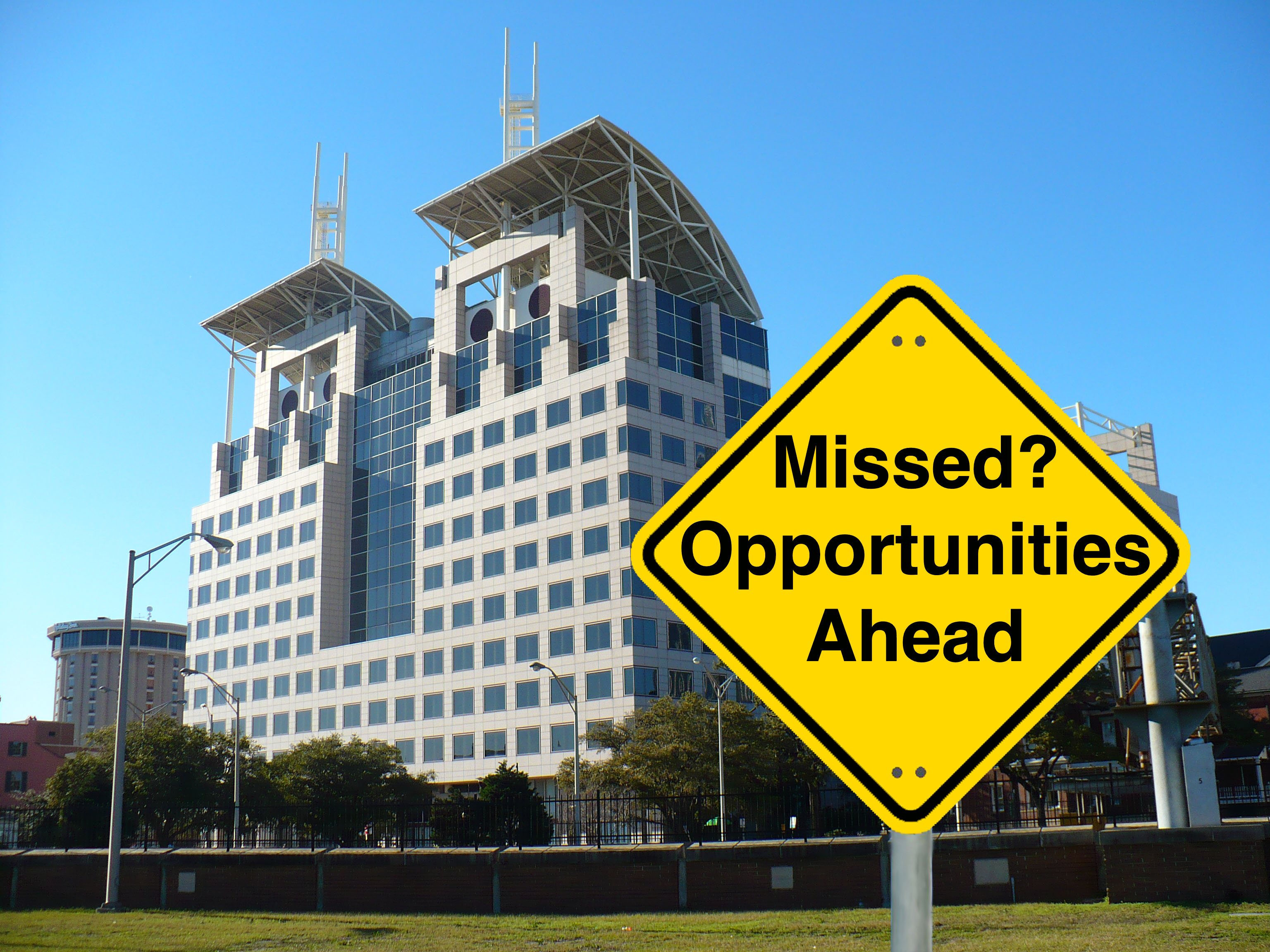 A yellow Caution road sign reading "Missed? Opportunities Ahead" in the foreground of a picture of Mobile Government Plaza